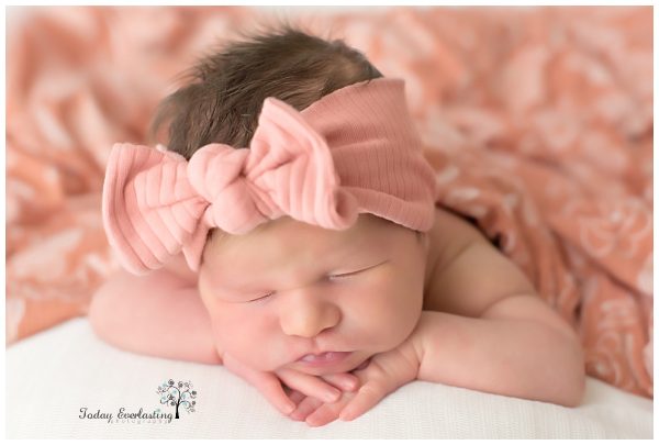 A peaceful newborn baby girl rests on her tummy, wearing a large, soft pink bow headband. Her tiny hands are tucked under her chin as she sleeps soundly on a cozy, peach-colored blanket, showcasing her delicate features and serene expression.