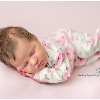 Newborn baby girl peacefully sleeping on a light pink backdrop, wearing a soft floral-patterned sleeper with pink flowers and green leaves. Her hands are gently tucked under her cheek, and her dark hair is slightly tousled. The photo is taken by Today Everlasting Photography, with the studio logo visible in the bottom right corner.