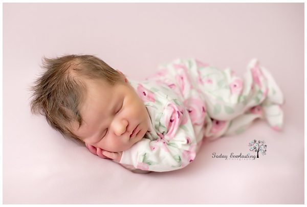 Newborn baby girl peacefully sleeping on a light pink backdrop, wearing a soft floral-patterned sleeper with pink flowers and green leaves. Her hands are gently tucked under her cheek, and her dark hair is slightly tousled. The photo is taken by Today Everlasting Photography, with the studio logo visible in the bottom right corner.