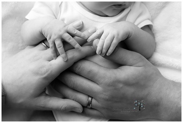 A black-and-white close-up of a newborn baby’s hands gently grasping both of her parents' fingers. The parents' hands, wearing wedding rings, cradle their baby's tiny hands, creating a tender moment of connection. The soft textures and delicate skin of the baby are captured beautifully, highlighting the love and bond within the family. The logo for Today Everlasting Photography is visible at the bottom of the image.